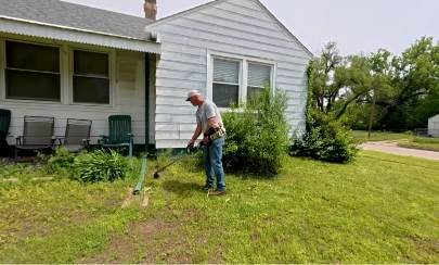 Lawn care technician trimming grass along a house foundation with a string trimmer